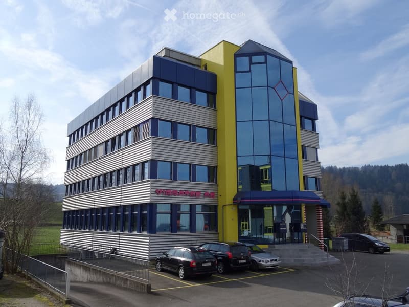 Office building with glass windows, yellow and blue sections, multiple cars parked in front