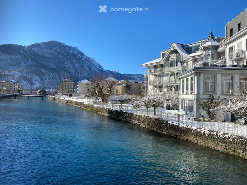 Riverfront of a town, buildings with snow on the rooftops, mountains in the background