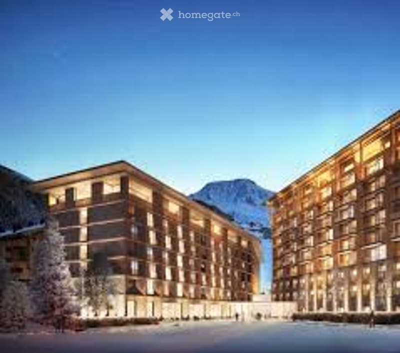 Modern hotel complex with multiple buildings, large windows, ground lighting, snowy landscape, mountain range in the background