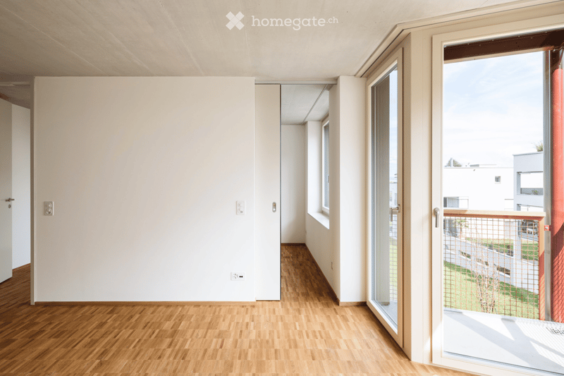 Empty room with wooden flooring, white walls, and large windows leading to a balcony area.