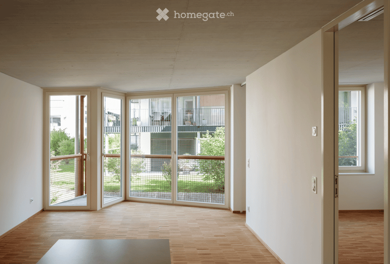 Bright, open room with light wooden flooring, white walls, and large sliding glass doors leading to a balcony with a view of greenery and neighboring buildings.