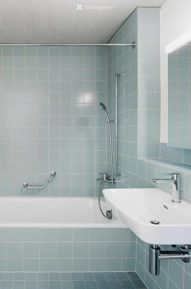 Modern bathroom with a white bathtub, a white sink, and a showerhead attached to a handheld hose. The walls and floor are tiled in light blue with white grout lines. A handrail is mounted on the wall for support.