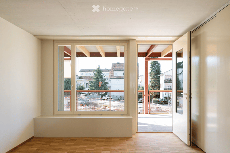Empty room with wooden flooring, large windows, and an open door leading to a balcony area. The room has white walls and a simple, modern design.