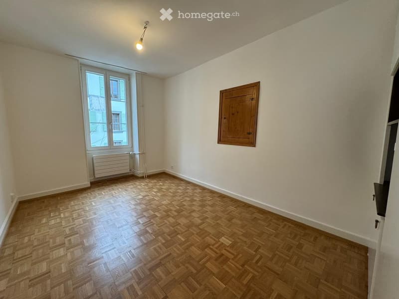Empty room, wooden parquet flooring, white walls, ceiling light, window with white frames, radiator, wooden wall frame, door