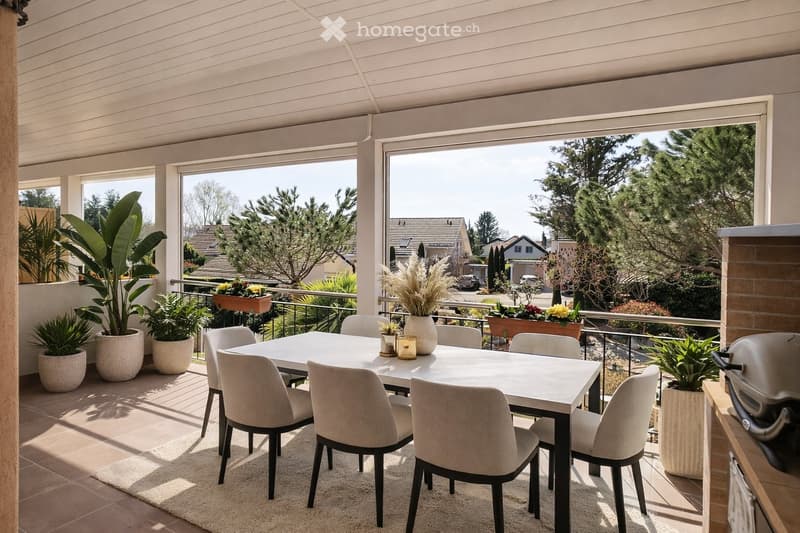 Balcony / Terrace. Balcony with a modern dining setup, including a white table, six chairs, a vase with dried flowers, and several potted plants. The balcony has a view of a suburban neighborhood with houses and trees.