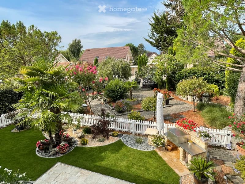 Backyard with palm tree, shrubs, flowers, patio, and grill; white picket fence surrounds; houses in the background; blue sky.