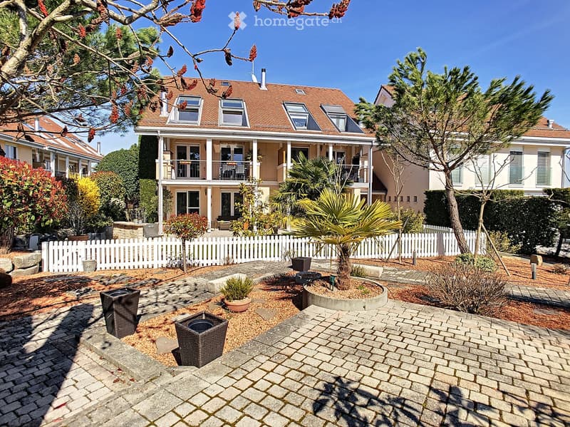 Three-story house, brown roof, beige walls, white fences, balconies, garden with brick walkway and potted plants, trees and shrubs