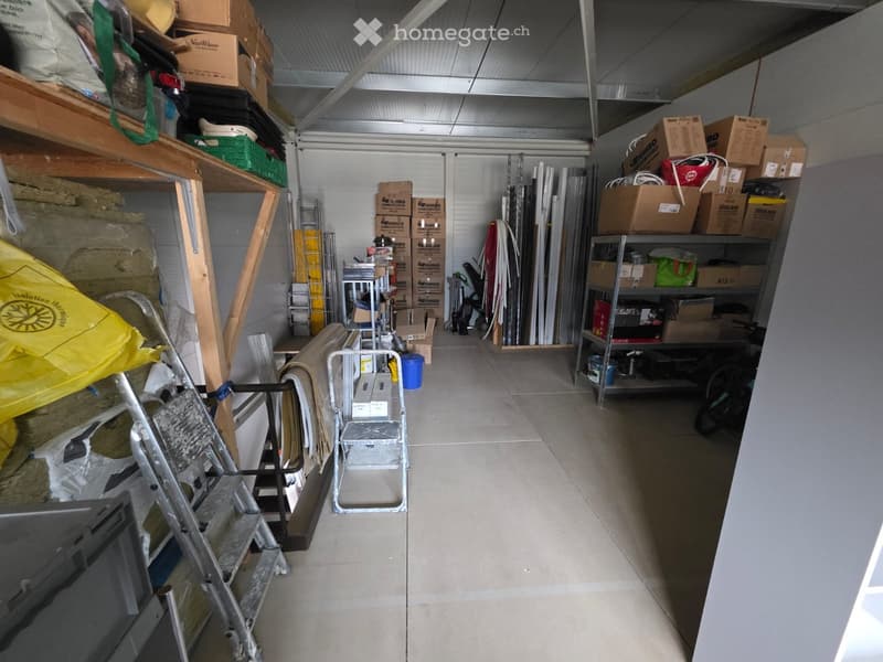 Storage room with shelves, boxes, ladders, a trolley, and some tools. The floor is concrete and appears to be empty. A bicycle is parked on the right side.