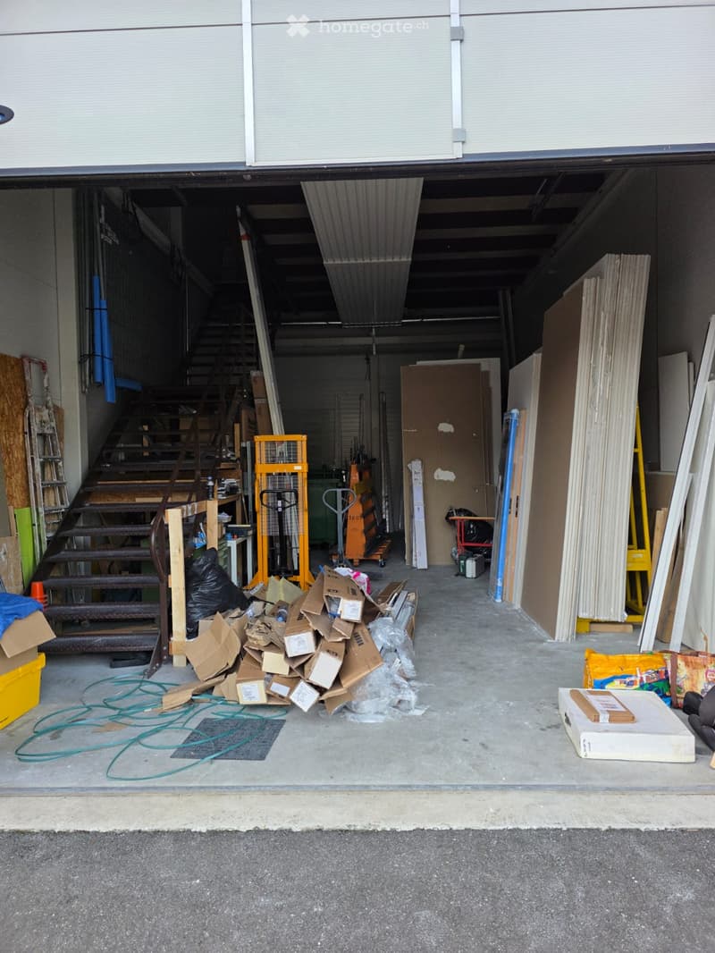 The garage appears to be cluttered with various boxes, building materials, and other miscellaneous items. The space has a staircase, storage boxes, wooden pallets, and a few trolleys.