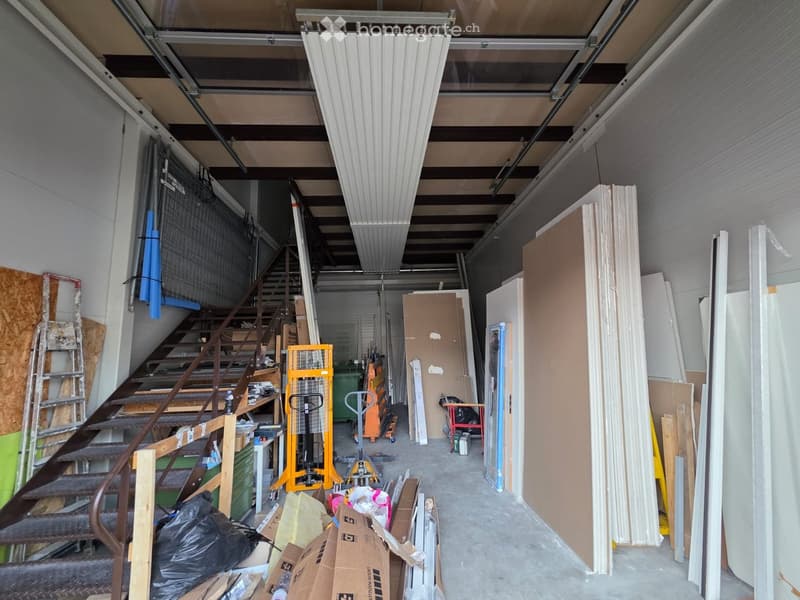 Empty garage with stairs, metal shelving, storage boxes, yellow machinery, ladders, trash bags, and cardboard boxes.