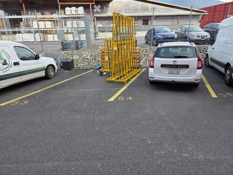outdoor parking lot with various cars and vans parked, yellow parking lines, yellow barriers, gravels, metal storage racks, building with a fenced area in the background