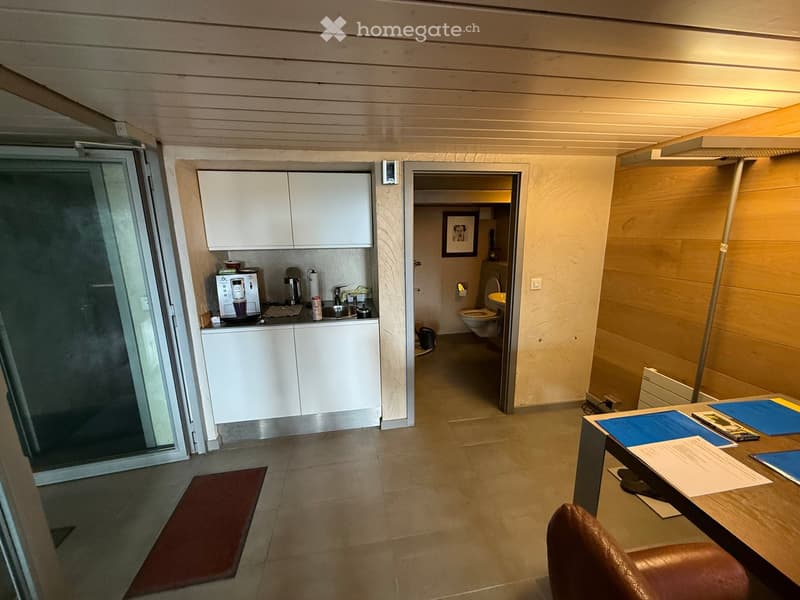 Compact kitchenette, sink, countertop, white cabinets, coffee machine, small table, chair, open door leading to a small bathroom, and a glass door leading to another room.