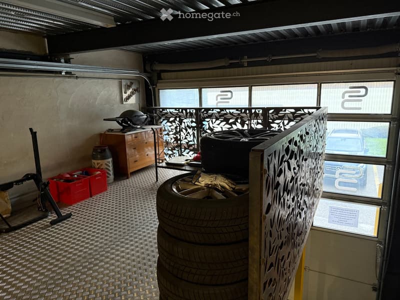 a small garage with a large door, a stack of tires, a car parked outside, a metal rack with a helmet, storage boxes, and a cabinet.