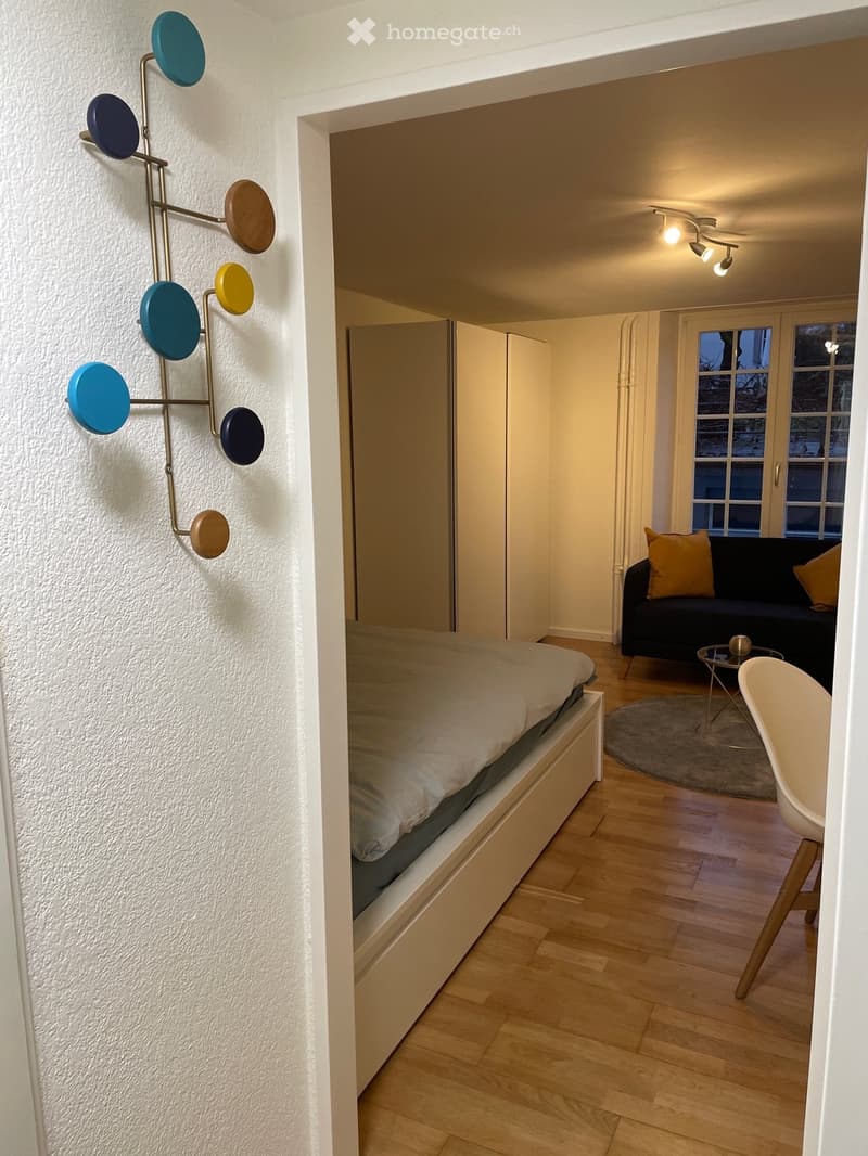 Small bedroom with a bed, coat rack, beige couch, and a side table.