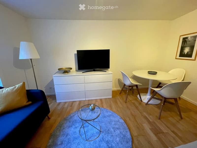 white walls, light wooden floors, yellow couch with pillow, glass side table, round glass top coffee table, white table with four chairs, white cabinet with television on top, floor lamp