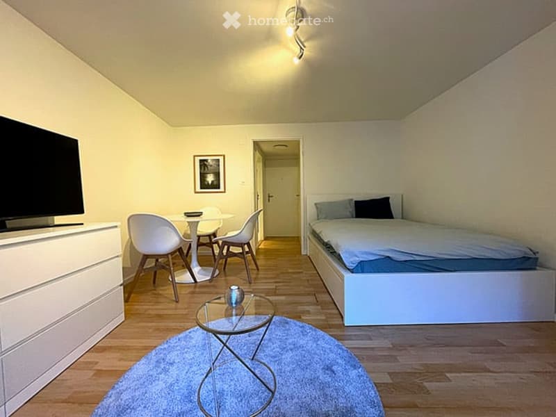 A small studio room with white walls, wooden floor, a single bed, a round table with 4 chairs, a glass center table, a TV and a white cabinet.