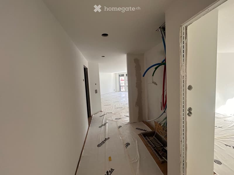 Empty hallway with white walls, tarpaulin on the floor, exposed pipes and wires, door on the right.