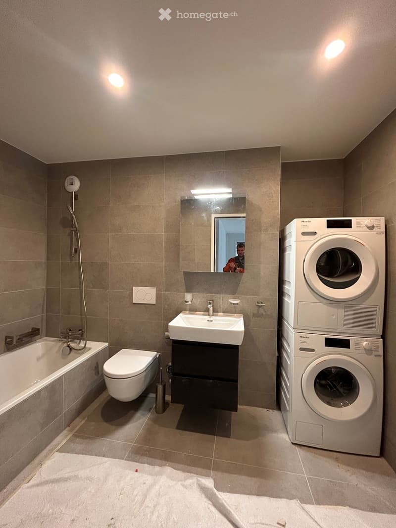 Modern bathroom with a bathtub, toilet, sink, mirror, and stacked washing machine/dryer unit.