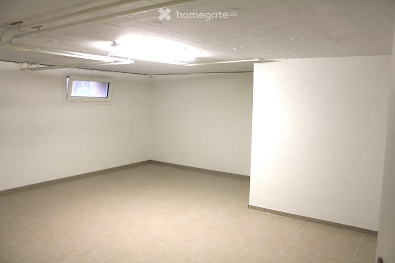 Empty room with white walls, tiled floor, small window, ceiling pipes, overhead light