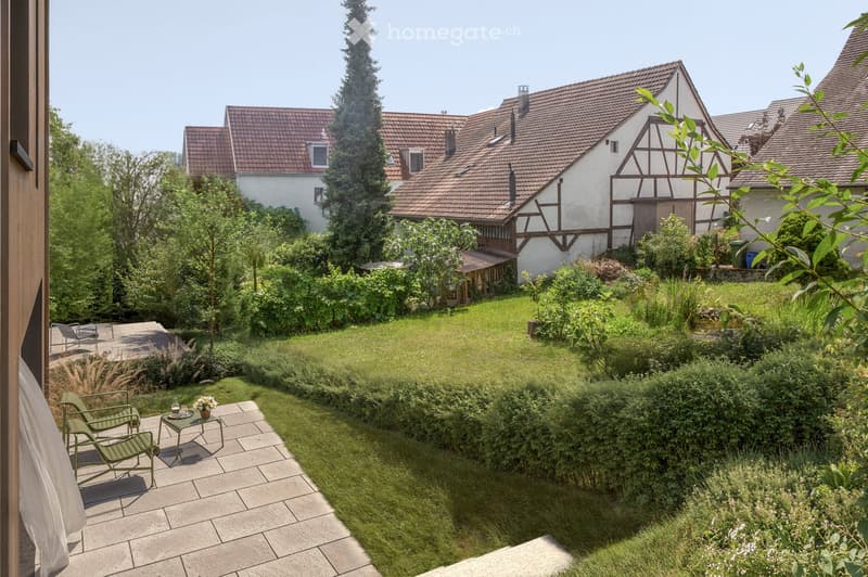 outdoor green space with patio, bushes, plants, green chairs, and a small table with a flower vase
