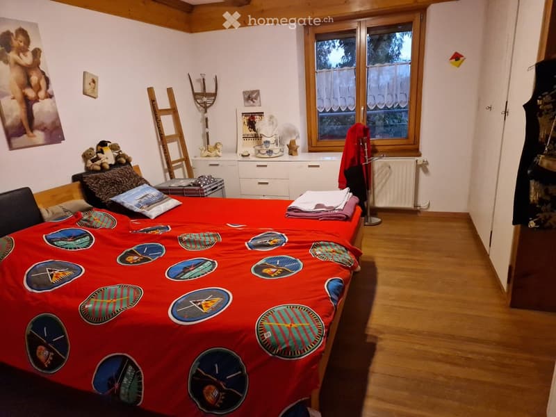 a bed with red bedsheets, decorative pillows, wooden floor, a painting of cherubs on the wall, a window with curtains, a radiator, and a cabinet under the window