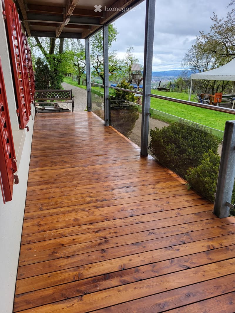 Wooden porch with red shutters, metal railings, large window, metal poles, outdoor bench, fenced area, trees and plants, grass, distant trees, view of the mountains