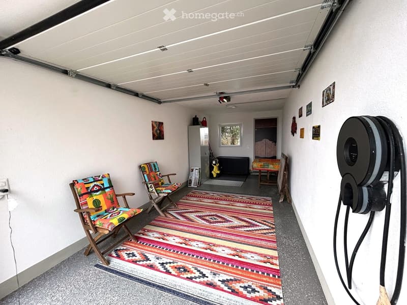 white walls, metal overhead garage door, hardwood floor, decorative rug, chairs, table, charger, storage box, potted plant