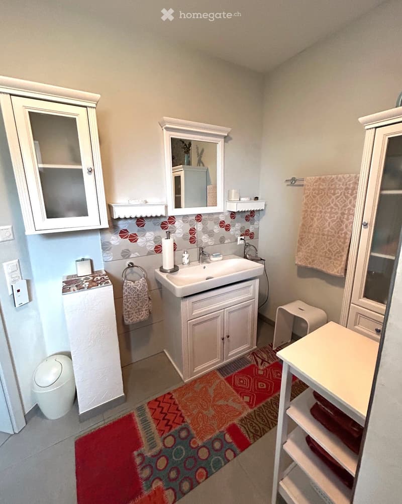 A bathroom with white tiled walls and floors, a white sink with a countertop, a white cabinet with a glass door, a mirror above the sink, a white trash can, and a small rug on the floor.