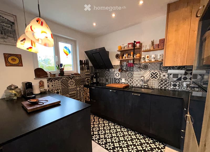 Black countertops, black cabinets, white tiled walls, black and white tiled floor, hanging lamps, coffee maker, sink, window, bowl and spoon on a wooden board