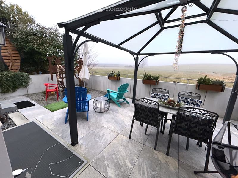 Gazebo, chairs, table, grassy area, concrete floor, potted plants, greenery, trees, view of landscape, white wall, black mat, gray patio