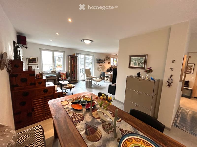 Spacious room with large windows, a wooden table with a decorative runner, and a dining set. Wooden cabinets are present along with framed pictures on the wall. A couch is partially visible.