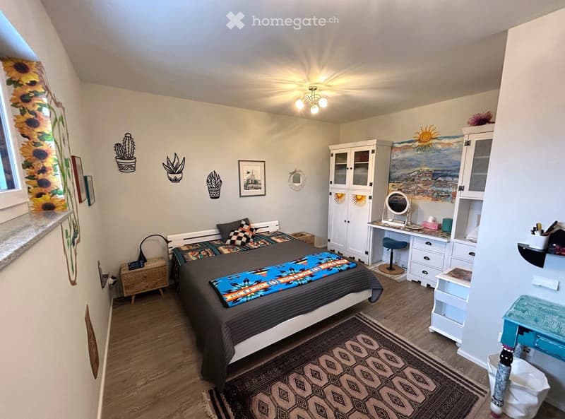 white wall, wooden floor, bed with blanket, desk with drawers, cabinets, lamp, artwork on wall, rug