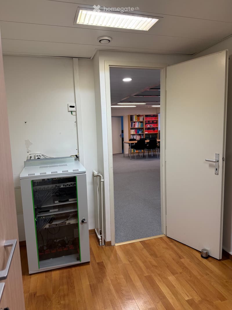 Office room, grey carpeted floor, white walls, open door, server rack, ceiling lights, radiator, small object in hallway