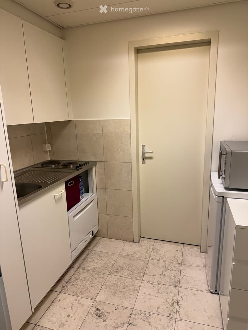 Modern and small kitchen with white cabinets, tiled flooring, a sink, a countertop with cooking plates, a dishwasher, and an electrical outlet. A white door leads to the adjoining room.