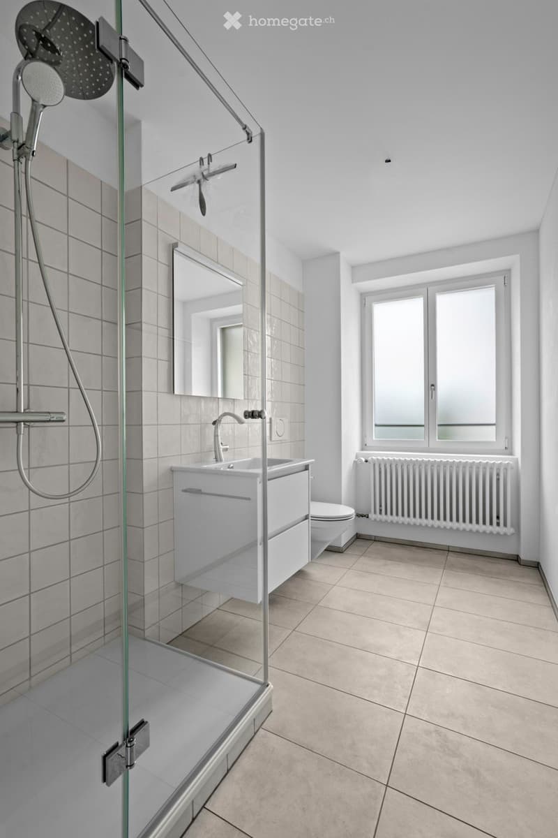 Modern bathroom with glass shower, white sink, white toilet, mirror, white tile floor, radiator