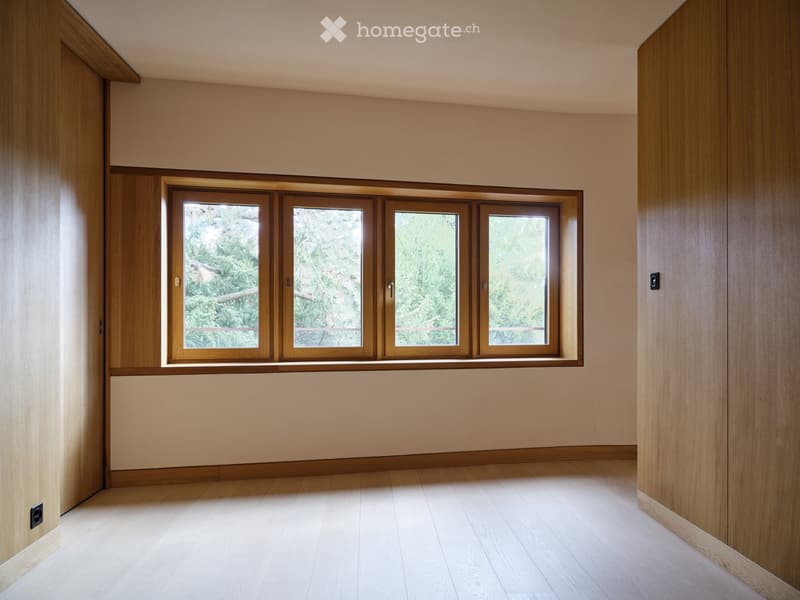 Empty room with light wooden flooring, beige walls, and large windows with wooden frames, offering a view of greenery outside.