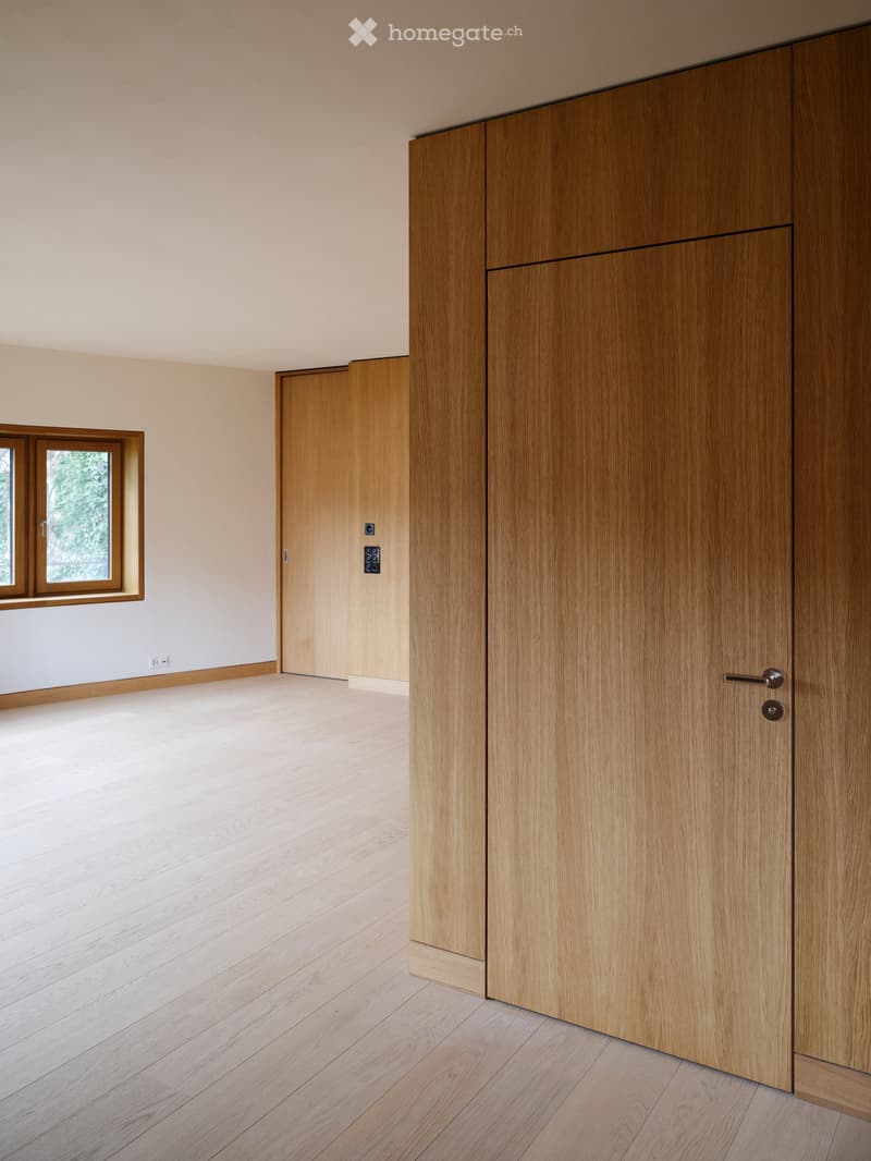 Empty room with light wooden flooring, light-colored walls, and a large window with wooden frame. Wooden doors are visible on the right side.