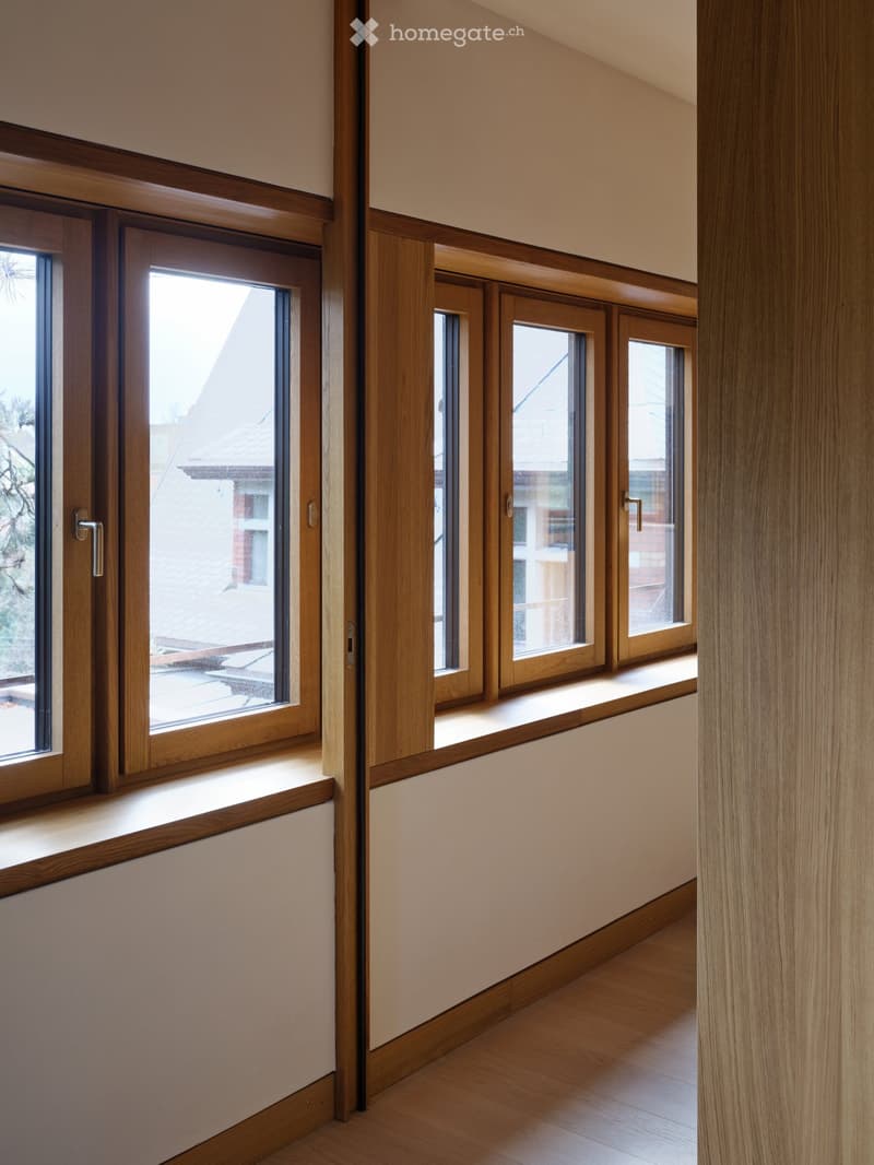 Bright hallway with wooden flooring, light-colored walls, and multiple large windows with wooden frames, offering natural light and a view of the outside.