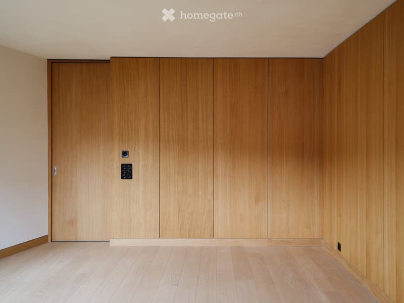 Empty room with light wooden flooring and matching wooden wall paneling, featuring a closed door and electrical outlets.