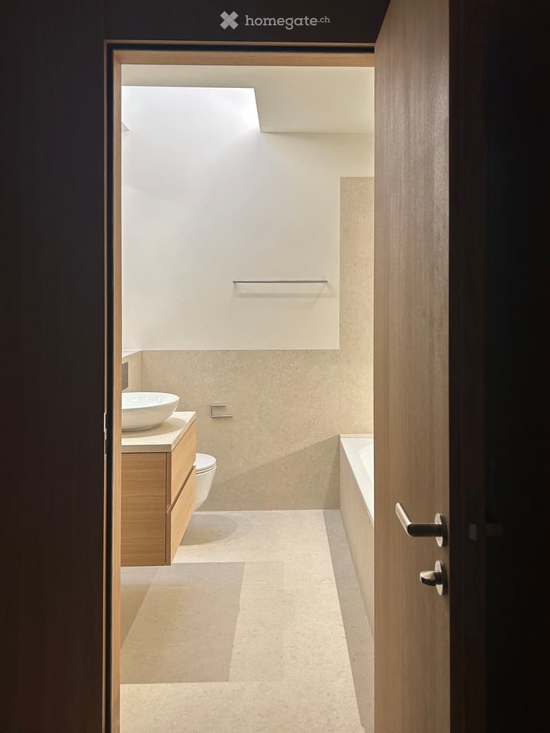 Modern bathroom with a sink, toilet, bathtub, and wooden cabinet. Light-colored tiles and a sleek design.