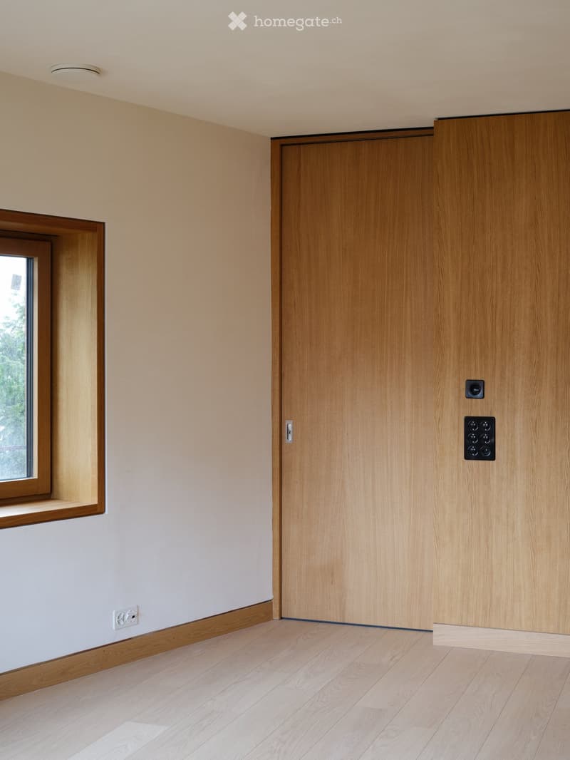 Empty room with light wooden flooring, white walls, a large window, and a wooden door.
