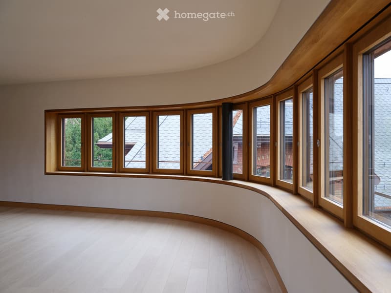 Empty room with curved walls, large wooden-framed windows, light wooden flooring, and a visible chimney pipe.