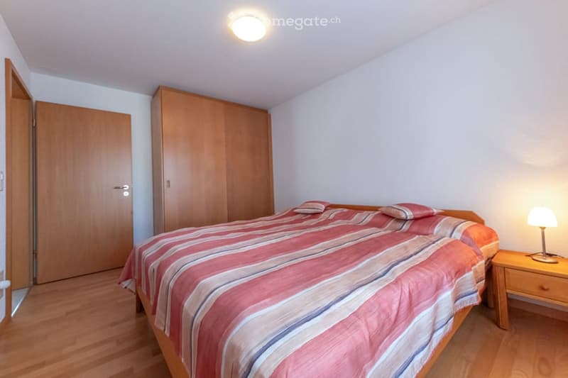 Bedroom with wooden floor, double bed, lamp on nightstand, wardrobe, and a white ceiling lamp.