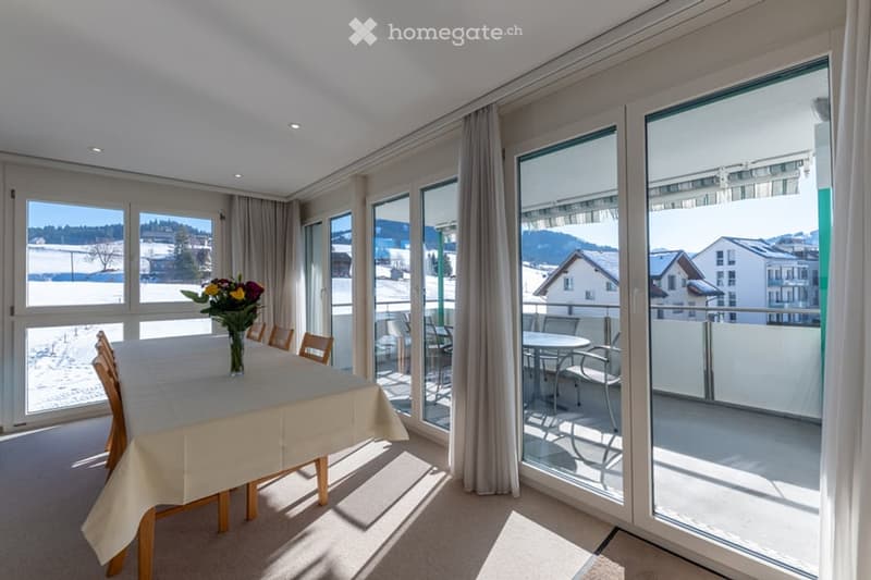 Dining room with large windows, dining table, chairs, and a vase with flowers. Sliding glass doors leading to a balcony with outdoor furniture. Mountain and snowy landscape view.