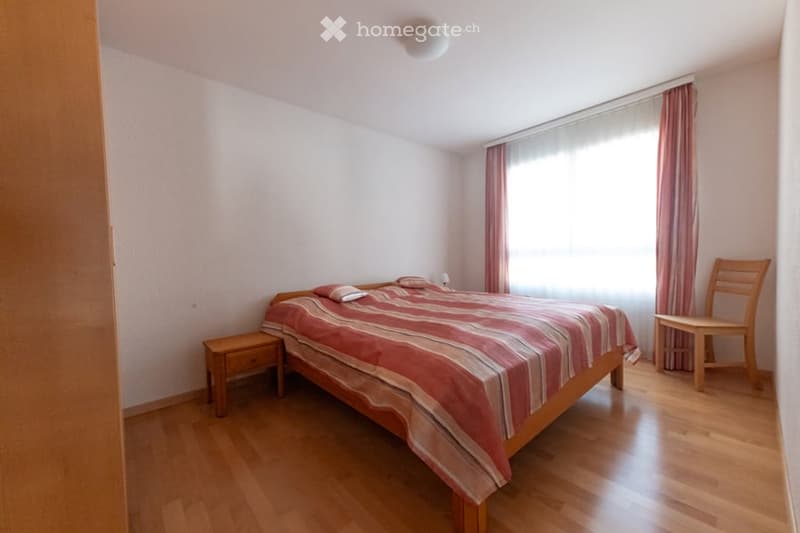 Bedroom with a double bed, wooden nightstand, wooden chair, white walls, wooden floor, lamp, large window with curtains