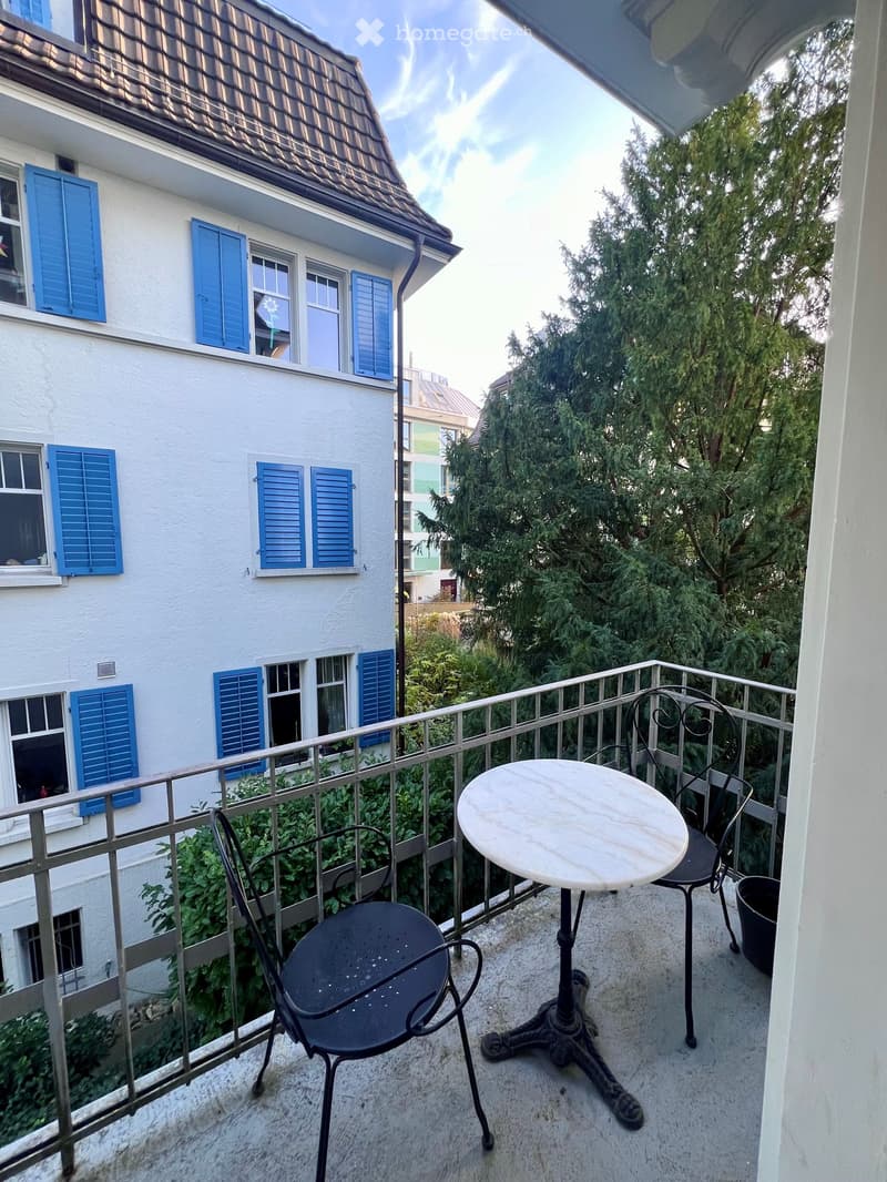 Marble table with two chairs, metal railings, garden view, green trees, white-washed wall, blue wooden shutters