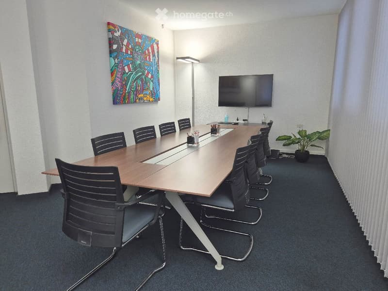 Long rectangular conference table, surrounded by black office chairs, white walls, mounted TV, potted plant, floor lamp.