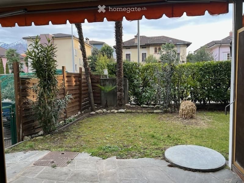 backyard, grassy area, gravel path, wooden fence, trees, bushes, round table, gate, mountains in the distance