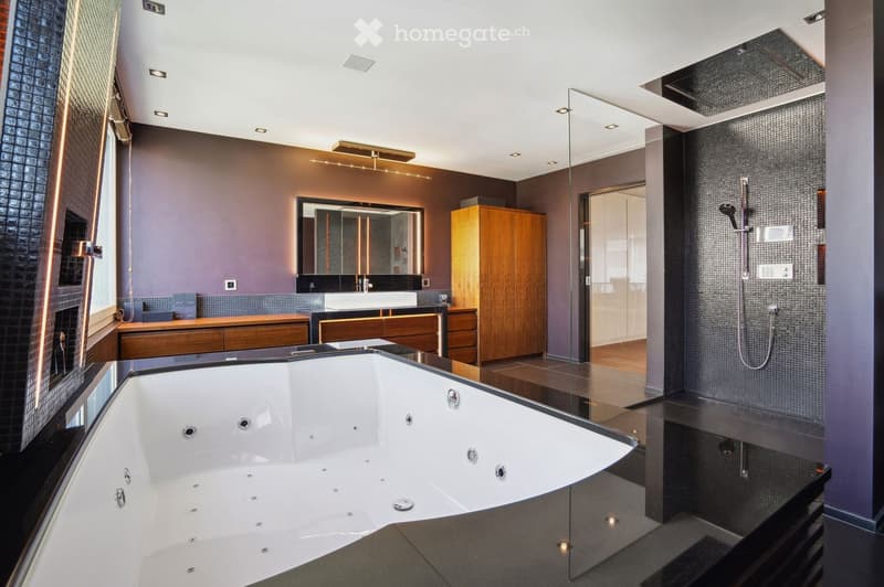 Large white bathtub with several jets, black granite top, wooden cabinets, dark tiles, glass wall, wooden flooring, mirror