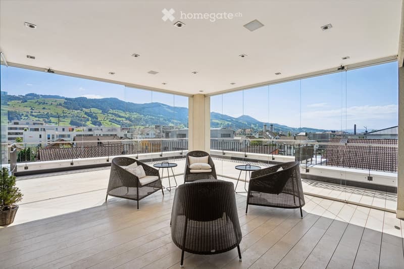 Wooden floor, wicker chairs, tables, decorative plant, railings, panoramic glass windows, mountain view